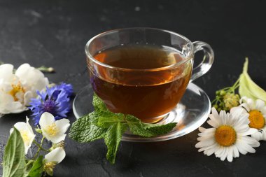 Cam fincanda aromatik bitki çayı, farklı çiçekler ve siyah masada nane şekeri, yakın plan.