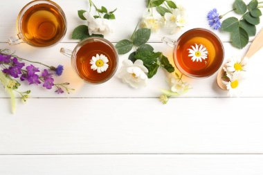Cam fincanlarda aromatik bitki çayı, farklı çiçekler ve beyaz ahşap masada yeşil yapraklar, düz yatak. Metin için boşluk