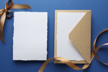 Düğün davetiyeleri, zarf, altın yüzükler ve mavi arka planda kurdele, düz yatış