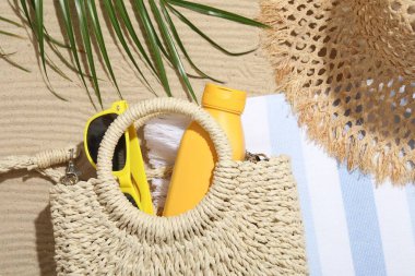 Plaj havlusu, hasır şapka, çanta, güneş kremi, güneş gözlüğü ve dışarıda kum üzerinde palmiye yaprağı.