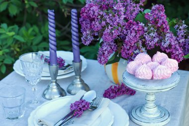 Güzel leylak çiçekleri, zephyrler, tabaklar, çatal bıçaklar, bardaklar ve mumlar bahçede, yakın plan