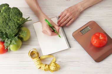 Hafif ahşap masada diyet planı geliştiren kadın mutfak tartısı, mezura ve sağlıklı yiyecekler, üst görünüm.