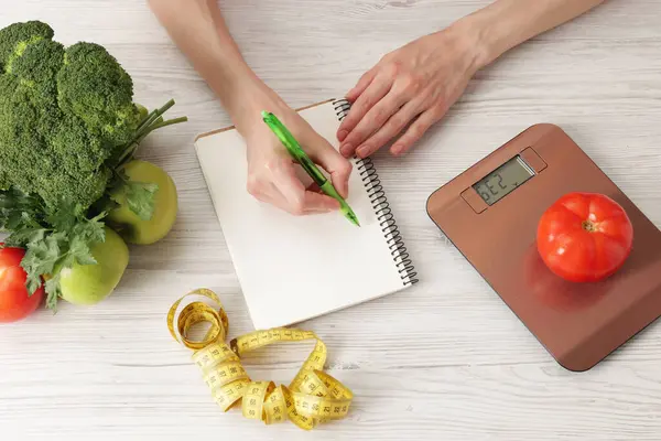 Hafif ahşap masada diyet planı geliştiren kadın mutfak tartısı, mezura ve sağlıklı yiyecekler, üst görünüm.