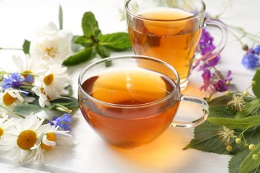 Cam bardaklarda Aromatik bitki çayı, farklı çiçekler ve beyaz ahşap masada yeşil yapraklar, yakın plan.