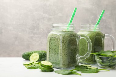 Mason kavanozlarındaki lezzetli smoothie, beyaz masada ıspanak yaprakları ve salatalık, gri arka planda, yakın planda.