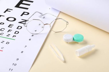 Görüş testi çizelgesi, gözlükler, cımbızlar, bej arka planda bir şişe tıbbi damla ve lensler, yakın plan.