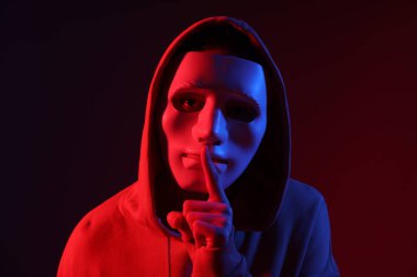 Anonimlik. Maske takan ve neon lambalı koyu kırmızı arka planda sessiz bir jest yapan bir kadın.