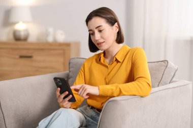 Evdeki koltukta modern akıllı telefon kullanan bir kadın.