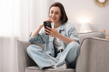 Evdeki koltukta modern akıllı telefon kullanan bir kadın.