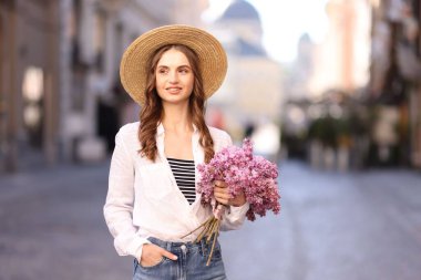 Şapkalı gülümseyen kadın, şehir caddesinde bir demet leylak çiçeğiyle.