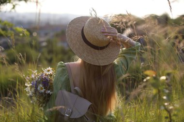 Güzel kır çiçekleriyle, şapkasıyla sırt çantasıyla güneşli bir günde çayırda gezen bir kadın.