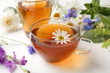 Cam bardaklarda Aromatik bitki çayı, farklı çiçekler ve beyaz ahşap masada yeşil yapraklar, yakın plan.