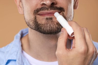 Açık kahverengi arka planda dudaklarına dudak kremi süren adam, yakın plan.