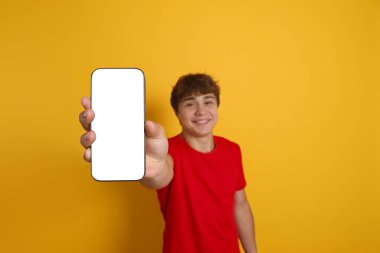 Gülümseyen genç çocuk sarı arka planda boş ekranlı akıllı telefon gösteriyor, seçici odaklanma. Tasarım için model