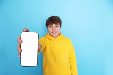 Gülümseyen genç çocuk açık mavi arka planda boş ekranlı akıllı telefon gösteriyor, seçici odaklanma. Tasarım için model