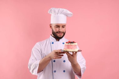 Üniformalı mutlu pastacı pembe arka planda böğürtlenli pasta tutuyor.