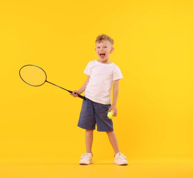 Sarı arka planda raketi ve tenis topu olan neşeli küçük bir çocuk.
