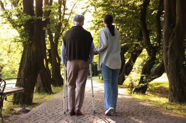 Bakıcı, dışarıda yürüyen çerçeve kullanan yaşlı bir adamı destekliyor.