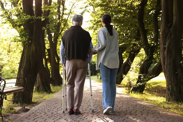 Bakıcı, dışarıda yürüyen çerçeve kullanan yaşlı bir adamı destekliyor.