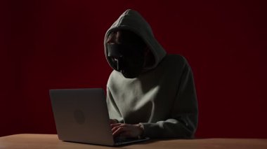 Anonimlik. Maske takan ve kırmızı arka plandaki tahta masada dizüstü bilgisayar kullanan bir kadın.