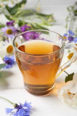 Cam bardakta Aromatik bitki çayı ve beyaz ahşap masada farklı çiçekler, yakın plan.