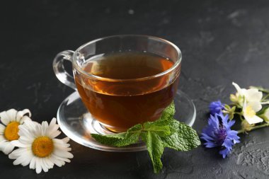 Cam fincanda aromatik bitki çayı, farklı çiçekler ve siyah masada nane şekeri, yakın plan.
