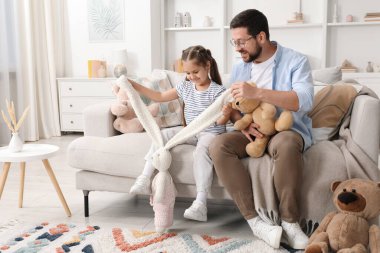 Baba ve kızı evdeki kanepede pelüş hayvanlarla oynuyorlar.