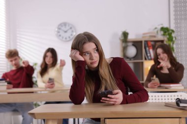 Siber zorbalık. Akıllı telefonlu ve sınıf arkadaşları tarafından sınıfa gösterilen, seçici odaklı üzgün bir genç kız.