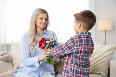 Evdeki kanepede gülümseyen annesine bir buket güzel çiçek sunan küçük çocuk.