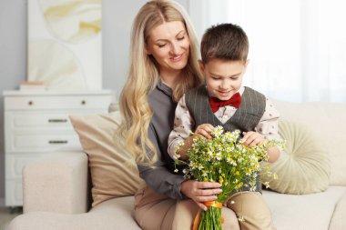 Küçük çocuk ve gülümseyen annesi evdeki kanepede bir buket güzel papatya çiçeğiyle