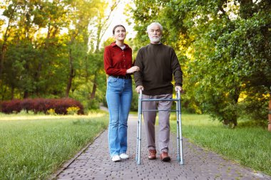 Gülümseyen bakıcı dışarıda yürüyen çerçeveyi kullanan yaşlı bir adamı destekliyor.
