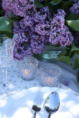 Güzel leylak çiçekleri, tabaklar, çatal bıçak takımı, cam ve yanan mumlar bahçede, yakın plan.