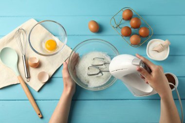 Bir kadın açık mavi ahşap masada el çırpıcı ile yumurta beyazı çırpıyor.