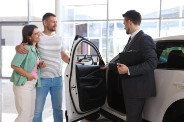 Elinde dosya olan satıcı salonda mutlu bir aileye yeni bir araba gösteriyor.