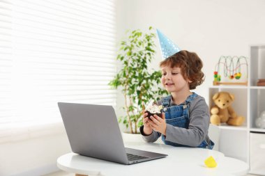 Doğum günü kutlaması. Elinde kek olan küçük çocuk içerideki masada dizüstü bilgisayarla video görüşmesi yapıyor. Metin için boşluk