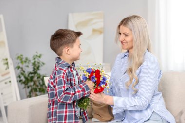 Evdeki kanepede gülümseyen annesine bir buket güzel çiçek sunan küçük çocuk.