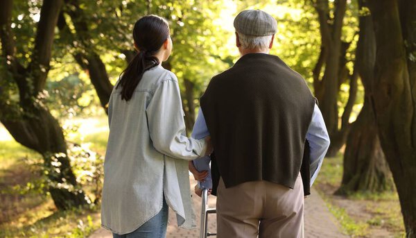 Caregiver supporting elderly man who using walking frame outdoors, back view