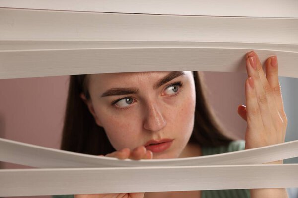 Paranoia. Worried woman looking through window blinds indoors
