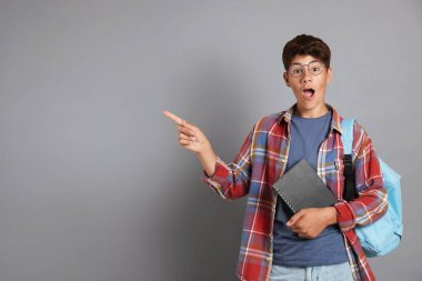 Not defteri ve sırt çantasıyla genç bir çocuk gri arka plandaki bir şeyi işaret ediyor, mesaj için boşluk.