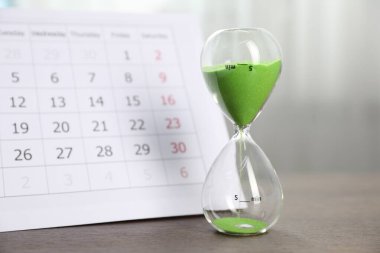 Son teslim tarihi. Kapalı alanda ahşap masada kum ve takvim akan kum saati, yakın plan.