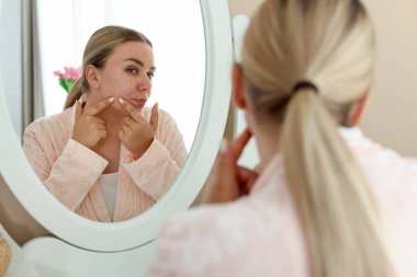 Evde aynaya bakan sivilceli bir kadın.