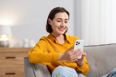 Evdeki koltukta modern akıllı telefon kullanan bir kadın.