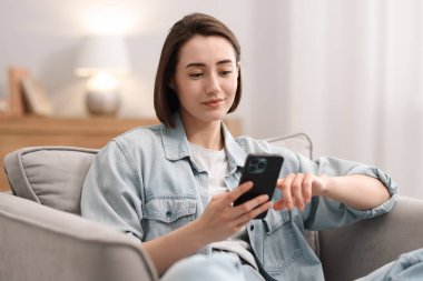 Evdeki koltukta modern akıllı telefon kullanan bir kadın.