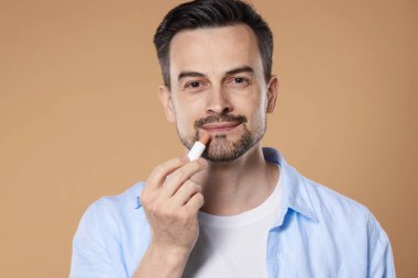 Yakışıklı bir adam açık kahverengi arka planda dudaklarına merhem sürüyor.
