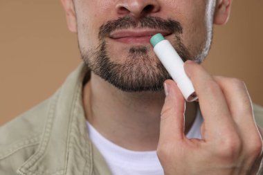 Açık kahverengi arka planda dudaklarına dudak kremi süren adam, yakın plan.