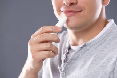 Gri arka planda dudaklarına dudak kremi süren genç adam, yakın plan.