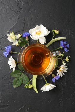 Cam bardakta Aromatik bitki çayı, farklı çiçekler ve siyah masada yeşil yapraklar, düz uzanmış