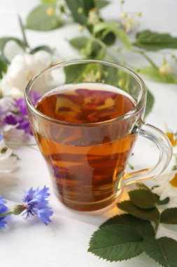 Cam bardakta Aromatik bitki çayı, farklı çiçekler ve beyaz ahşap masada yeşil yapraklar, yakın plan.