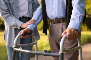 Bakıcı, dışarıda yürüyen çerçeve kullanan yaşlı bir adamı destekliyor.