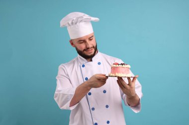 Üniformalı mutlu pastacı elinde böğürtlen ve açık mavi arka planda lezzetli pastalar tutuyor.